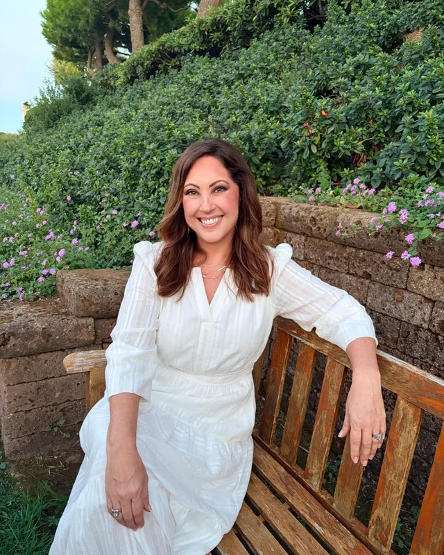 Jaime Estorge in a white dress, smiling on a wooden garden bench